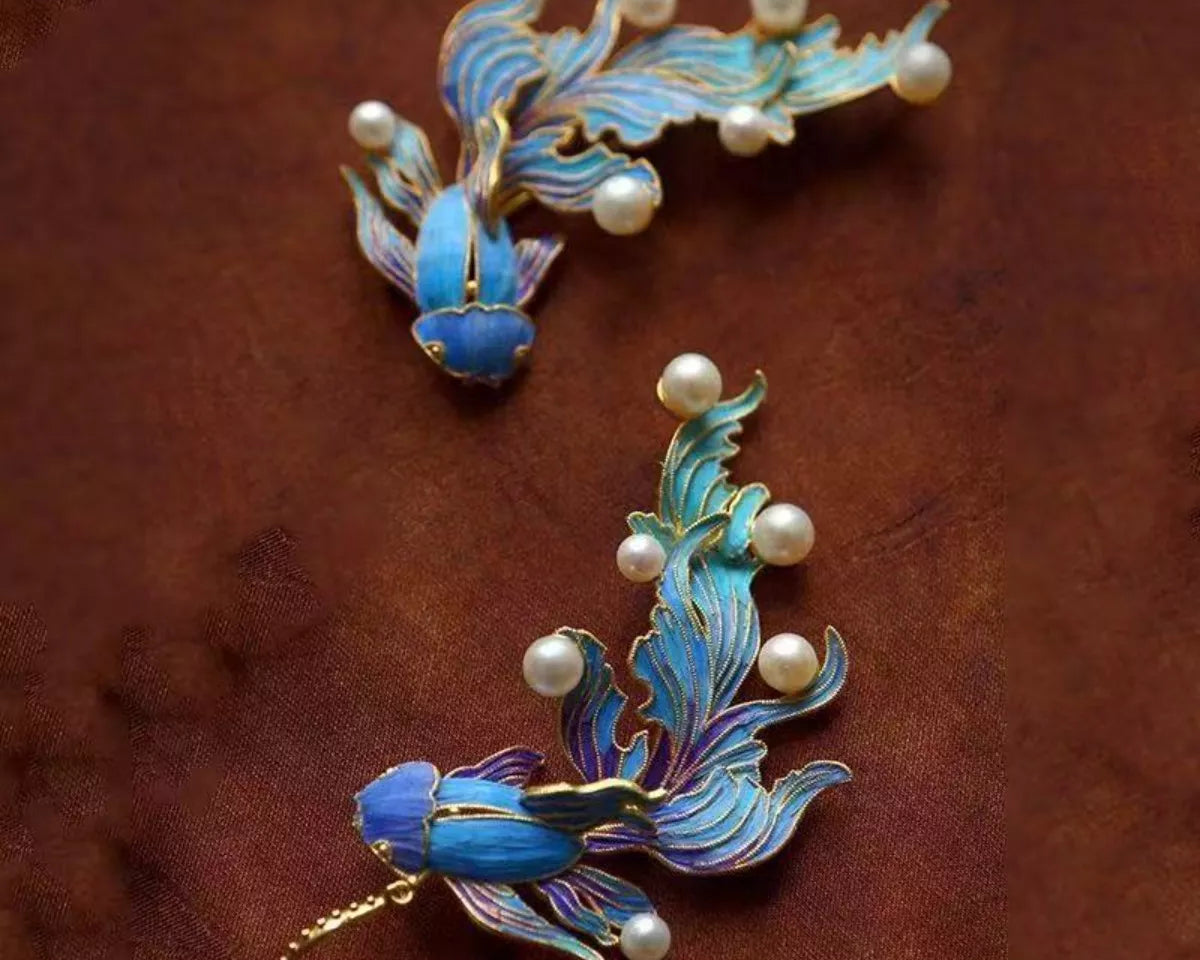 NYLD GLAMOUR By NEWYORKLISTED: Pair of blue fish earrings with gold accents and pearl-like beads on a brown background