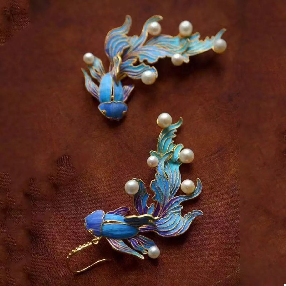 NYLD GLAMOUR By NEWYORKLISTED: Pair of blue fish earrings with gold accents and pearl-like beads on a brown background