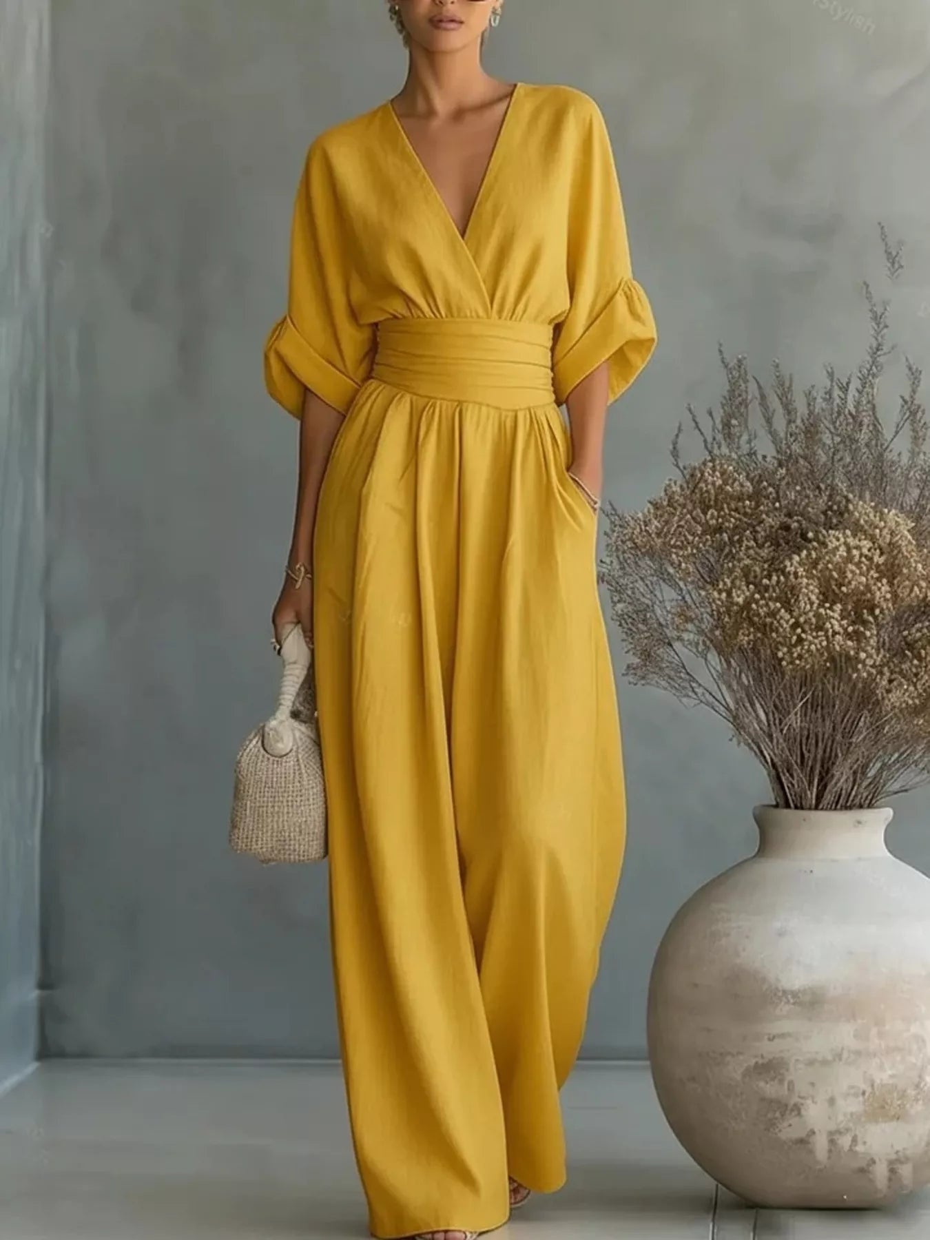 NEWYORKLISTED STYLE COLLECTION: Woman wearing a yellow jumpsuit standing next to a vase with dried flowers against a gray wall.