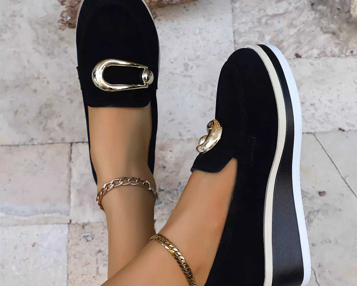 NEWYORKLISTED SHOE COLLECTION: Black slip-on shoes with gold accents worn by a person on a light stone floor.