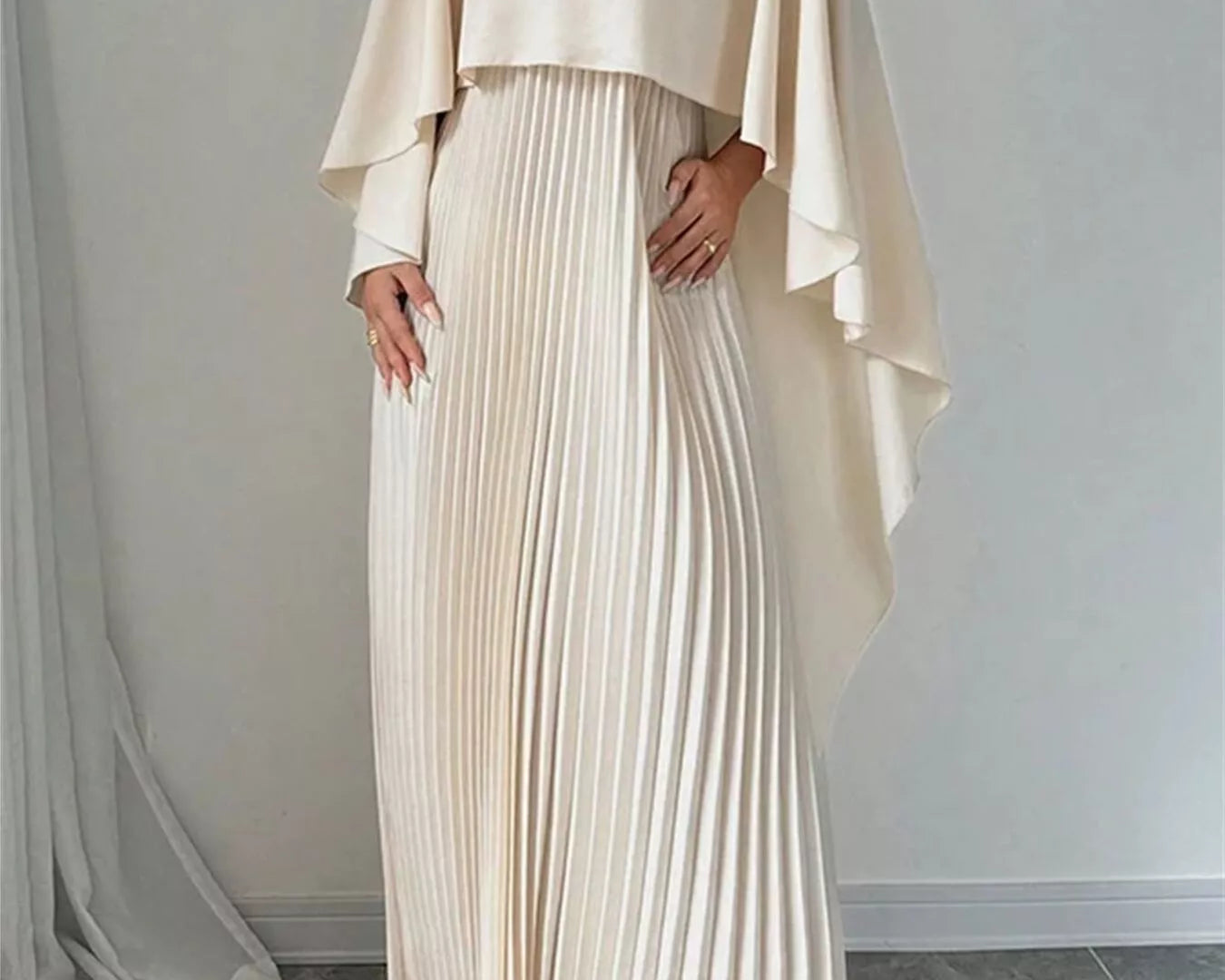 NEWYORKLISTED STYLE COLLECTION: Woman wearing a beige pleated dress with a matching cape against a plain background