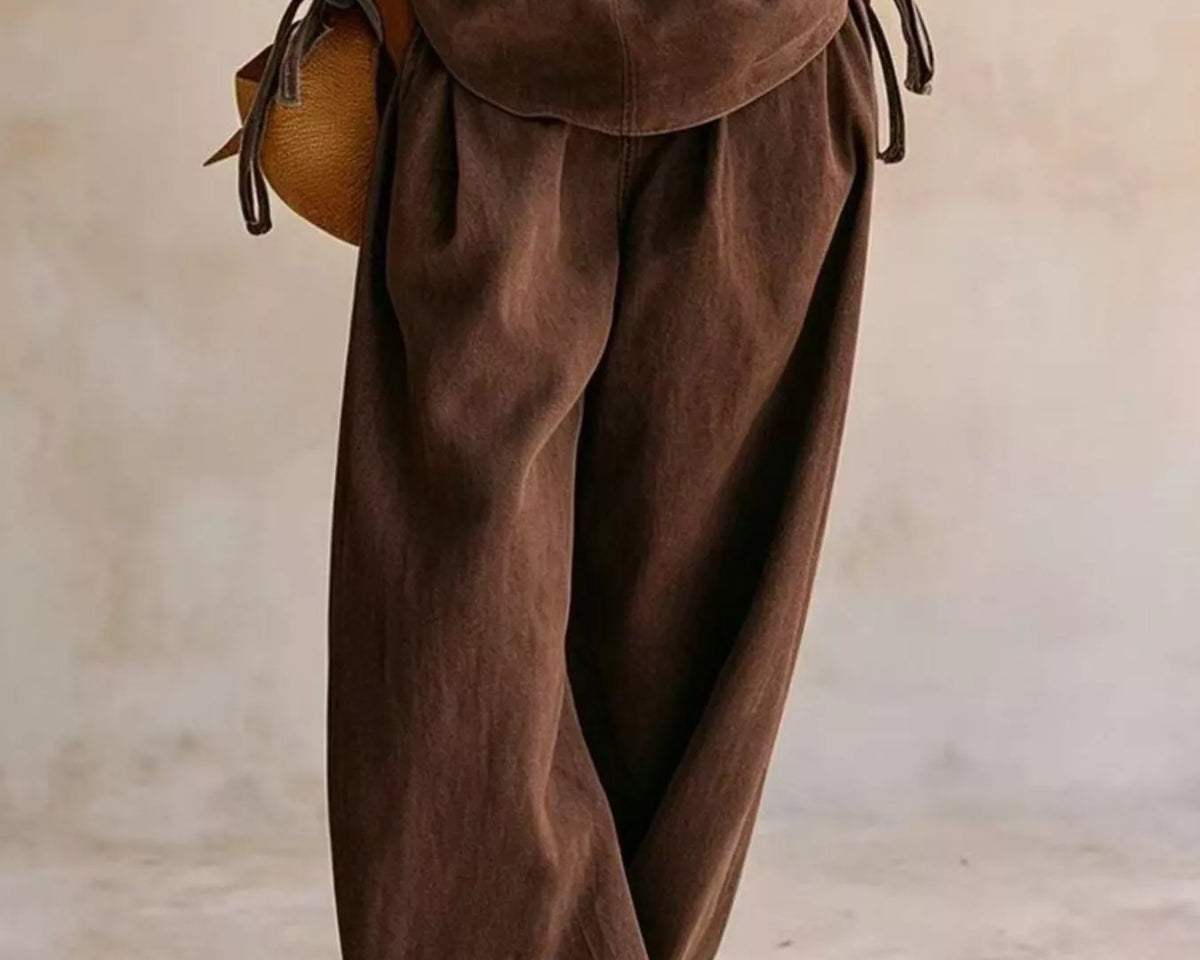 NEWYORKLISTED STYLE COLLECTION: A person wearing a matching brown linen-like two-piece outfit with a long-sleeve top and very loose wide-leg pants, standing against a neutral beige wall. A small tan woven bag is also visible.