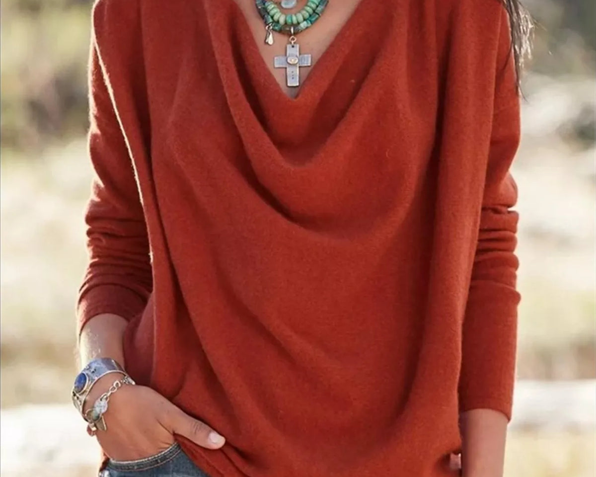 NEWYORKLISTED STYLE COLLECTION: A woman wearing a rust-orange long-sleeve knit top with a draped cowl neckline, paired with blue jeans and layered necklaces, photographed outdoors against a blurred natural background.