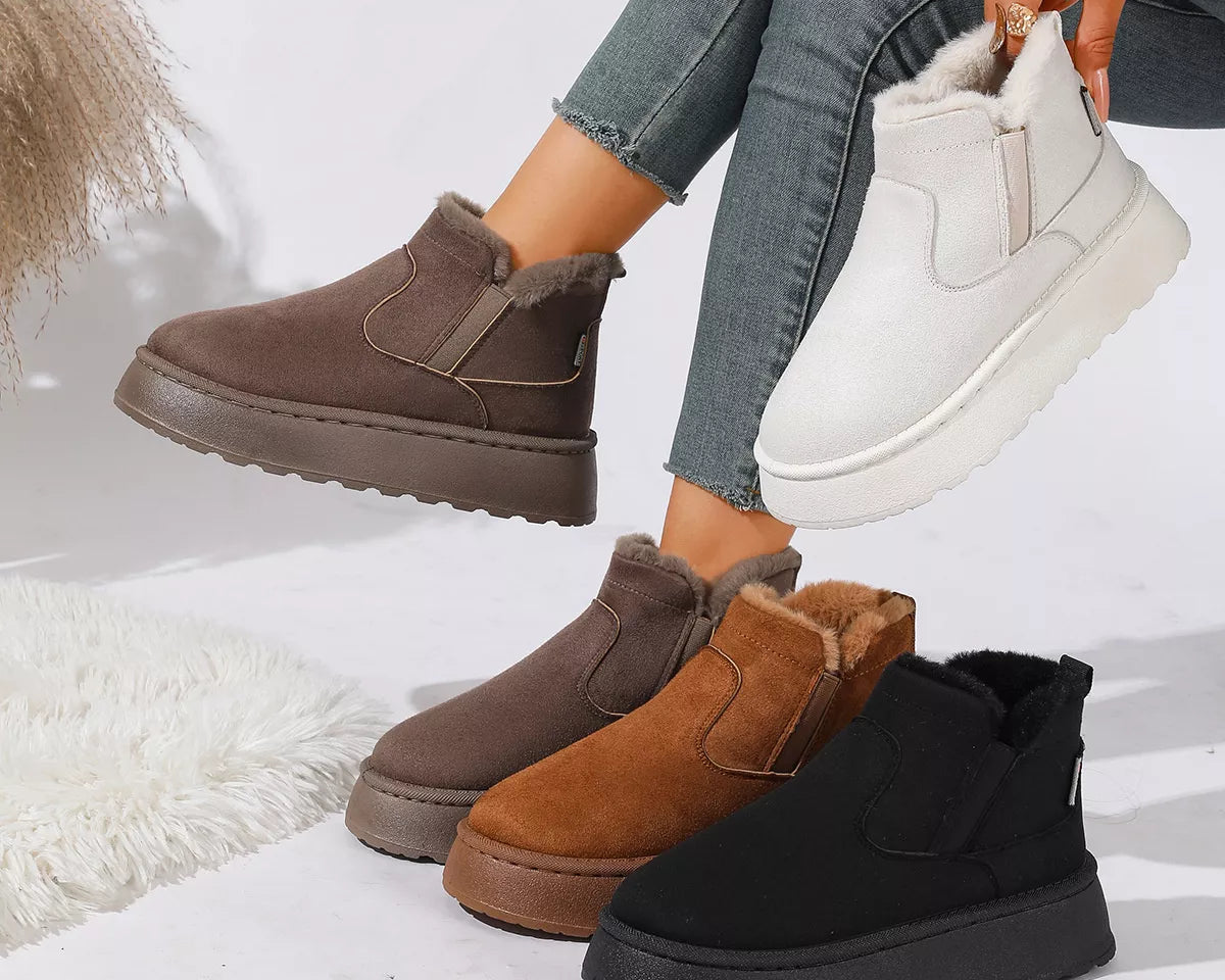 NEWYORKLISTED SHOE COLLECTION: Four slip-on platform ankle boots in brown, tan, black, and white are displayed on a white studio floor, with a person's legs and hand holding one of the white boots.