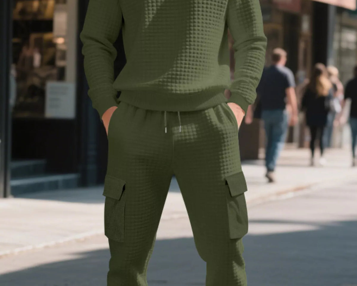 NEWYORKLISTED ATLAS MEN'S COLLECTION: A man wearing an olive green waffle-knit crewneck sweatshirt and matching cargo jogger pants, standing on a city sidewalk.