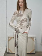 NEWYORKLISTED STYLE COLLECTION: Woman wearing a long-sleeve top with intricate patterns standing against a plain wall.