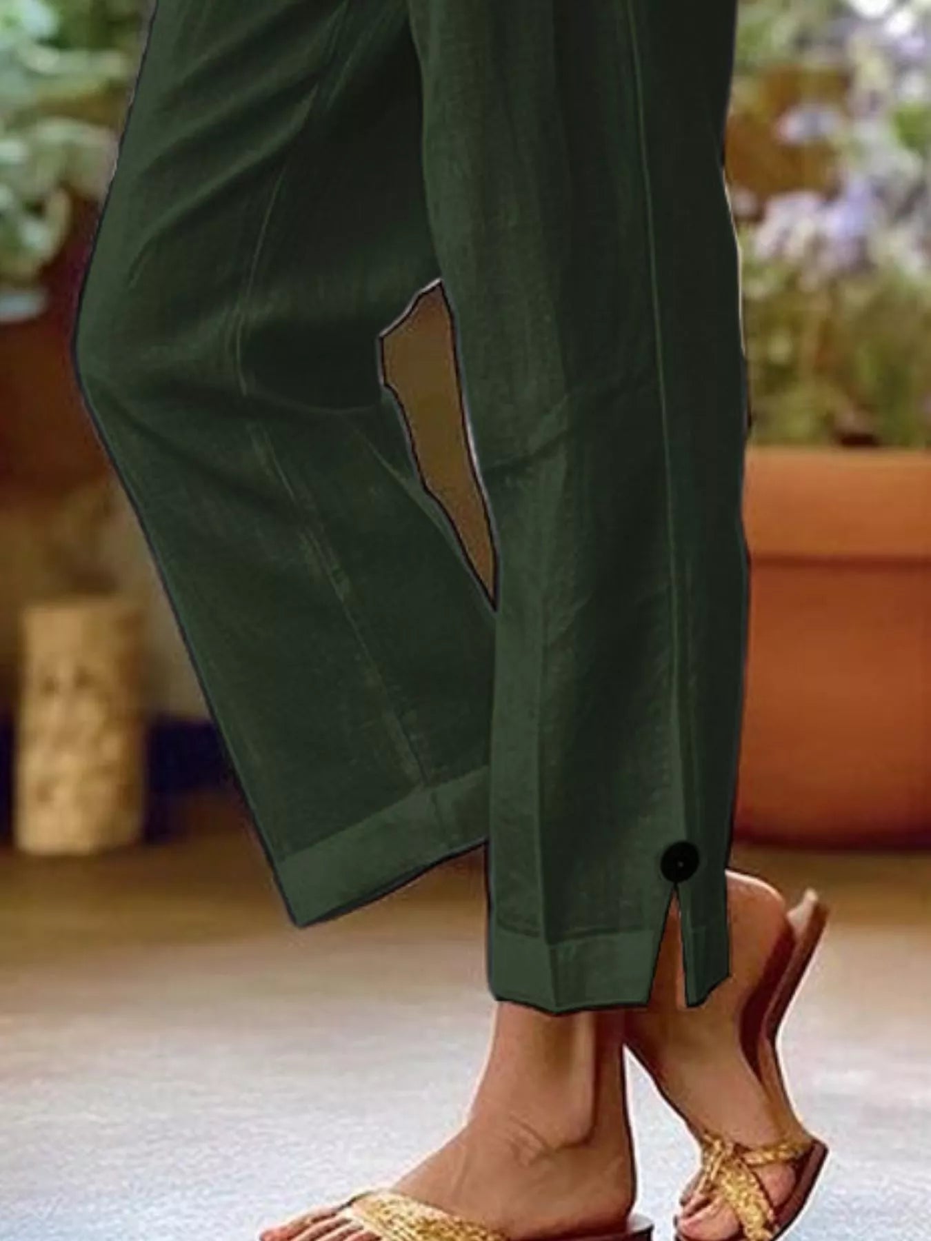 NEWYORKLISTED STYLE COLLECTION: A close-up of a person wearing dark olive green wide-leg pants with a side slit and a small black button detail near the hem, paired with gold-toned slide sandals.