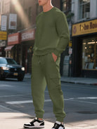 NEWYORKLISTED ATLAS MEN'S COLLECTION: A man wearing a matching olive green textured knit sweatshirt and jogger pants set, standing on a city street with storefronts and a car in the background.