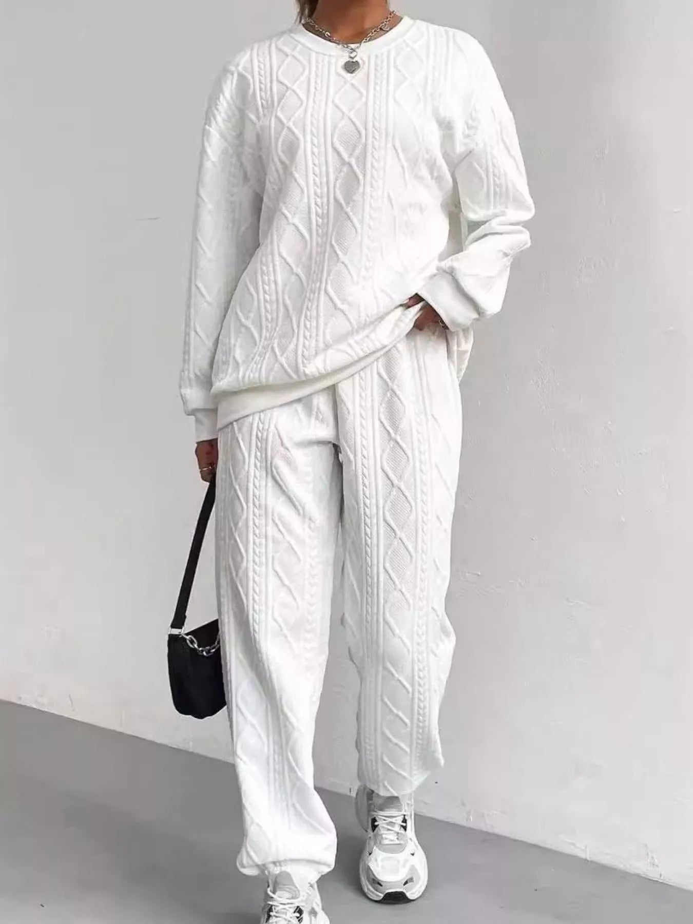 NEWYORKLISTED COMFORT COLLECTION: Person wearing a white cable knit sweater and pants with sneakers, holding a black bag against a light gray background.