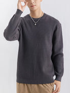 NEWYORKLISTED ATLAS MEN'S COLLECTION: A man wearing a dark gray ribbed knit crewneck sweater, paired with tan pants and a silver necklace, against a plain light gray background.