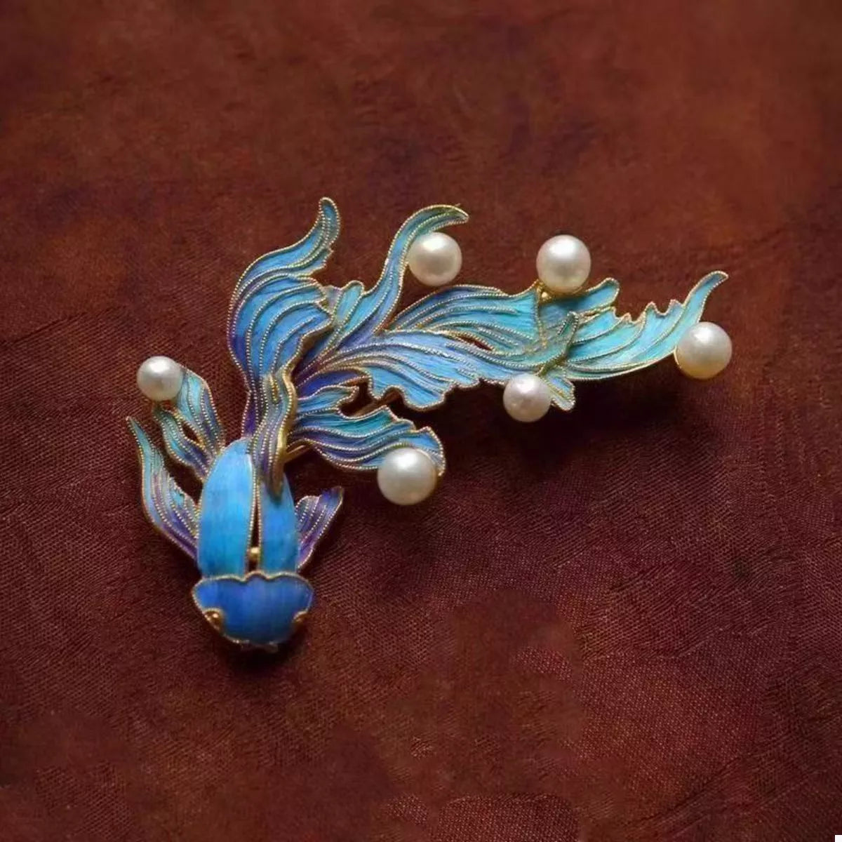 NYLD GLAMOUR By NEWYORKLISTED: Jewelry brooch with blue fish design and pearl-like beads on a brown fabric background