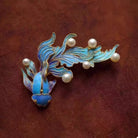 NYLD GLAMOUR By NEWYORKLISTED: Jewelry brooch with blue fish design and pearl-like beads on a brown fabric background