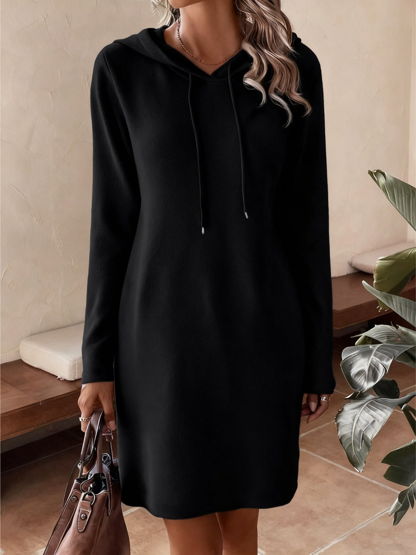 NEWYORKLISTED Comfort Collection: Person wearing a black hoodie dress holding a handbag indoors.