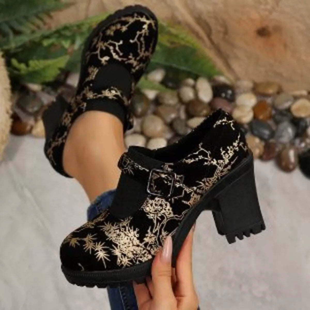 NEWYORKLISTED: Black velvet shoes with gold floral patterns held by a person against a natural background.