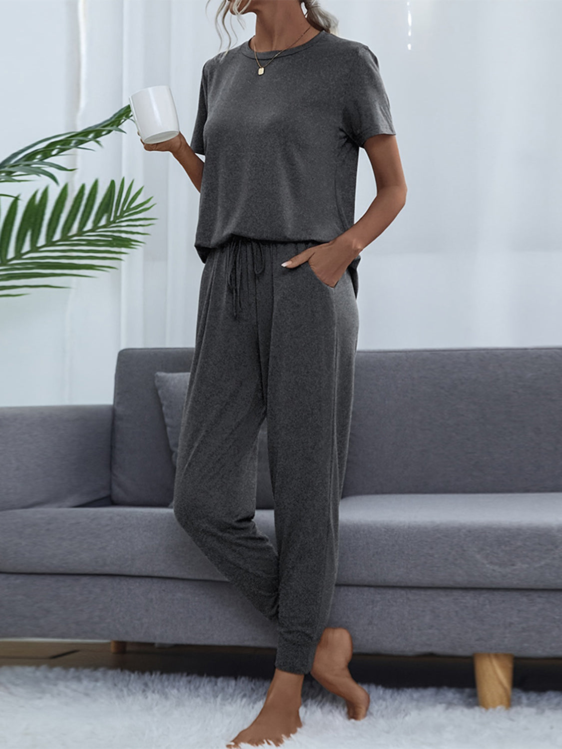NEWYORKLISTED Intimate Apparel: Person wearing a gray lounge set sitting on a gray couch with a white wall and plant in the background.