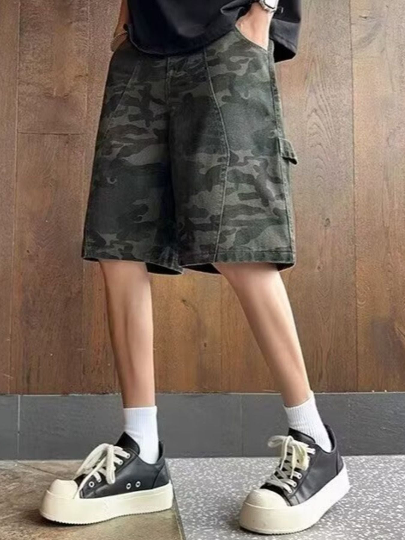 NEWYORKLISTED ATLAS MEN's COLLECTION: Person wearing camouflage shorts and black sneakers against a wooden wall.