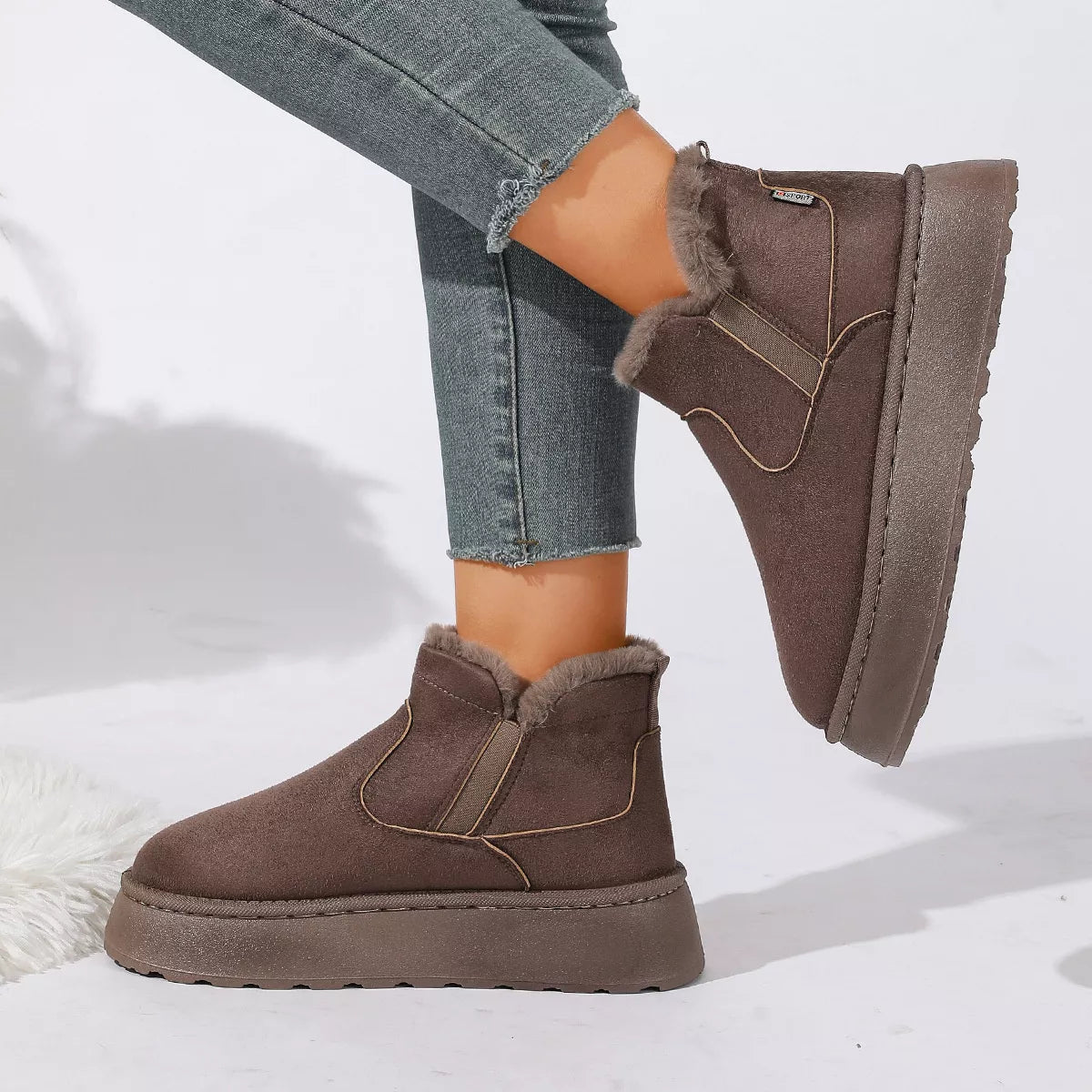 NEWYORKLISTED SHOE COLLECTION: A pair of women's mid-calf slip-on boots in taupe brown faux suede with a fuzzy lining, styled with frayed blue jeans on a white studio background.