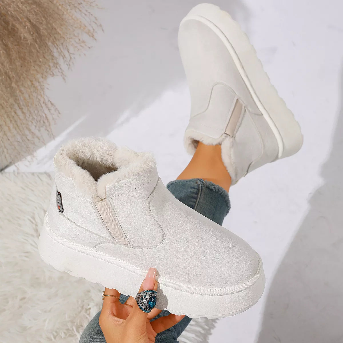 NEWYORKLISTED SHOE COLLECTION: A pair of off-white winter slip-on boots with plush faux-fur lining, shown worn with blue jeans and held by a hand against a bright, modern background.