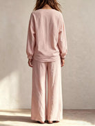 NEWYORKLISTED STYLE COLLECTION: A woman viewed from behind wearing a light pink linen two-piece set consisting of a long-sleeve top with gathered cuffs and matching wide-leg pants, standing against a textured beige wall.