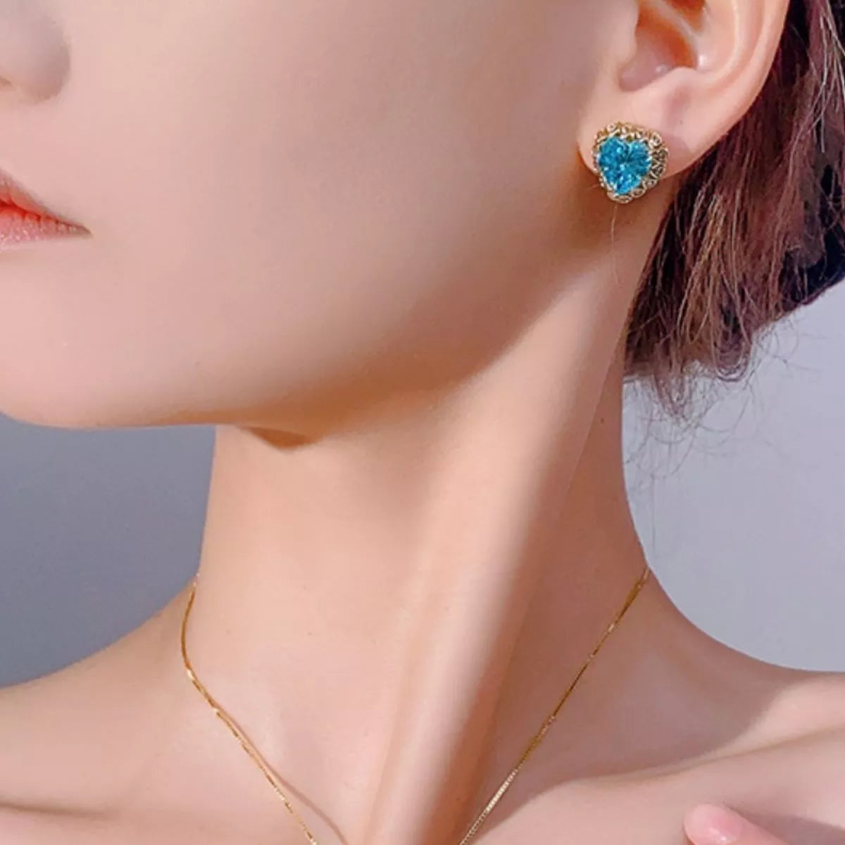 NYLD GLAMOUR COLLECTION By NEWYORKLISTED: Close-up of a woman wearing blue heart-shaped earrings with a blurred background