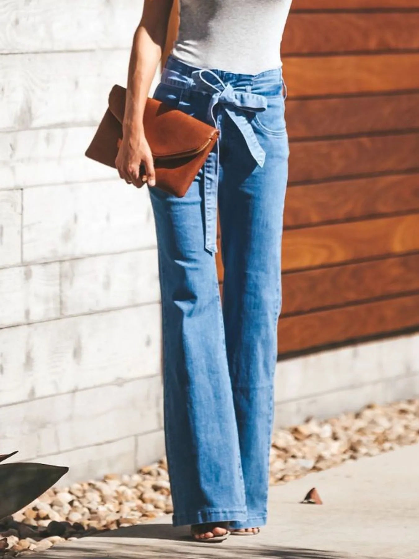 NEWYORKLISTED STYLE COLLECTION: Person wearing blue jeans and a white top, holding a brown clutch against a wooden wall.