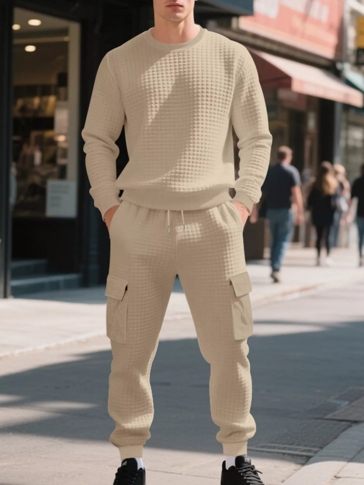 NEWYORKLISTED ATLAS MEN'S COLLECTION: A man wearing a matching beige waffle-knit sweatshirt and cargo jogger pants stands on a city sidewalk, with blurred storefronts and pedestrians in the background.