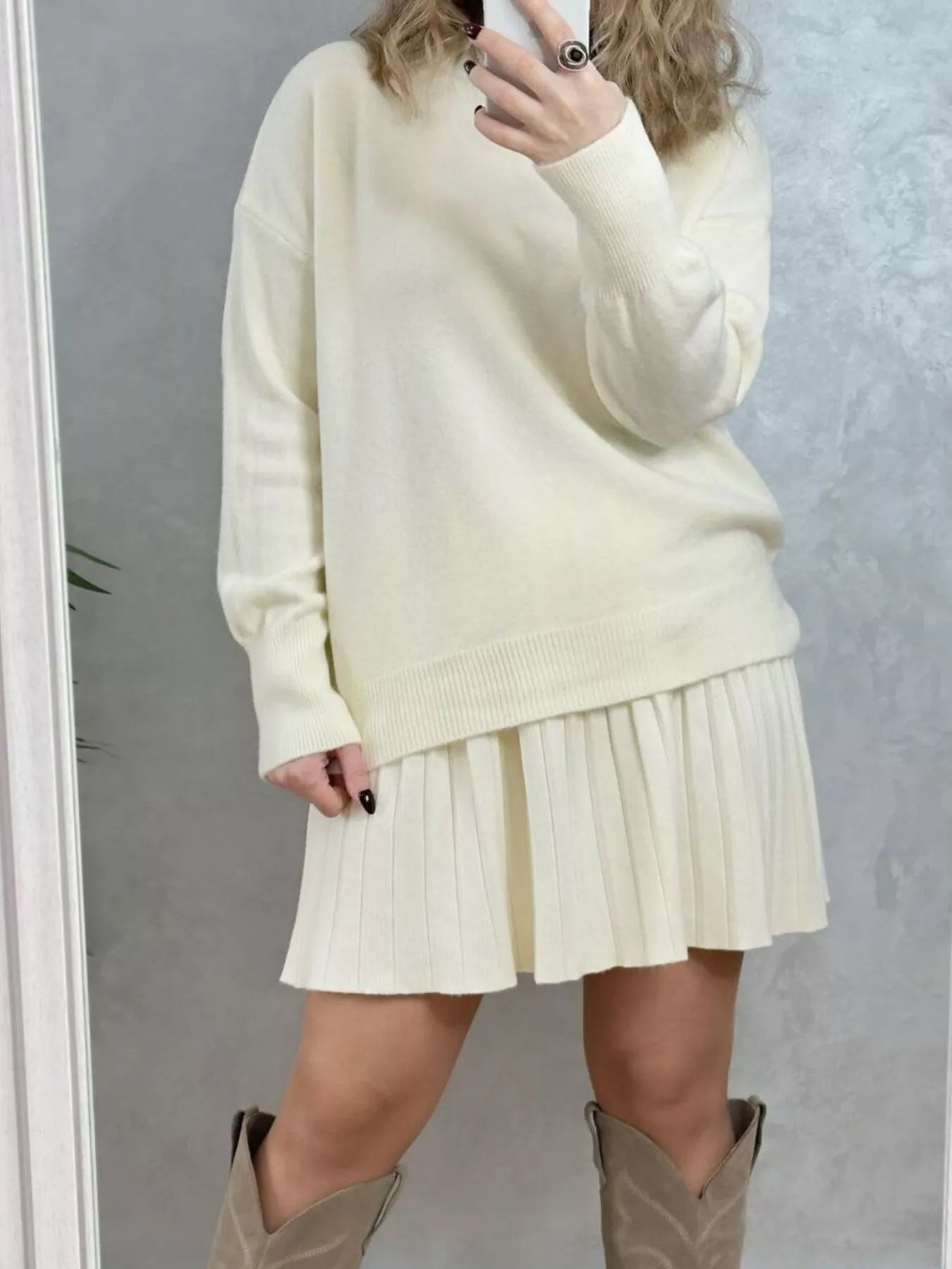 Person wearing a cream-colored sweater and pleated skirt with brown boots against a plain background on NEWYORKLISTED.COM