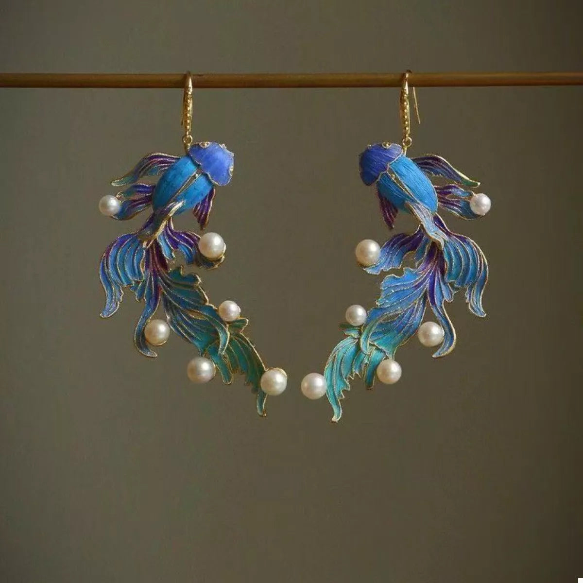 NYLD GLAMOUR By NEWYORKLISTED: Pair of blue fish-shaped earrings with pearl accents on a dark background