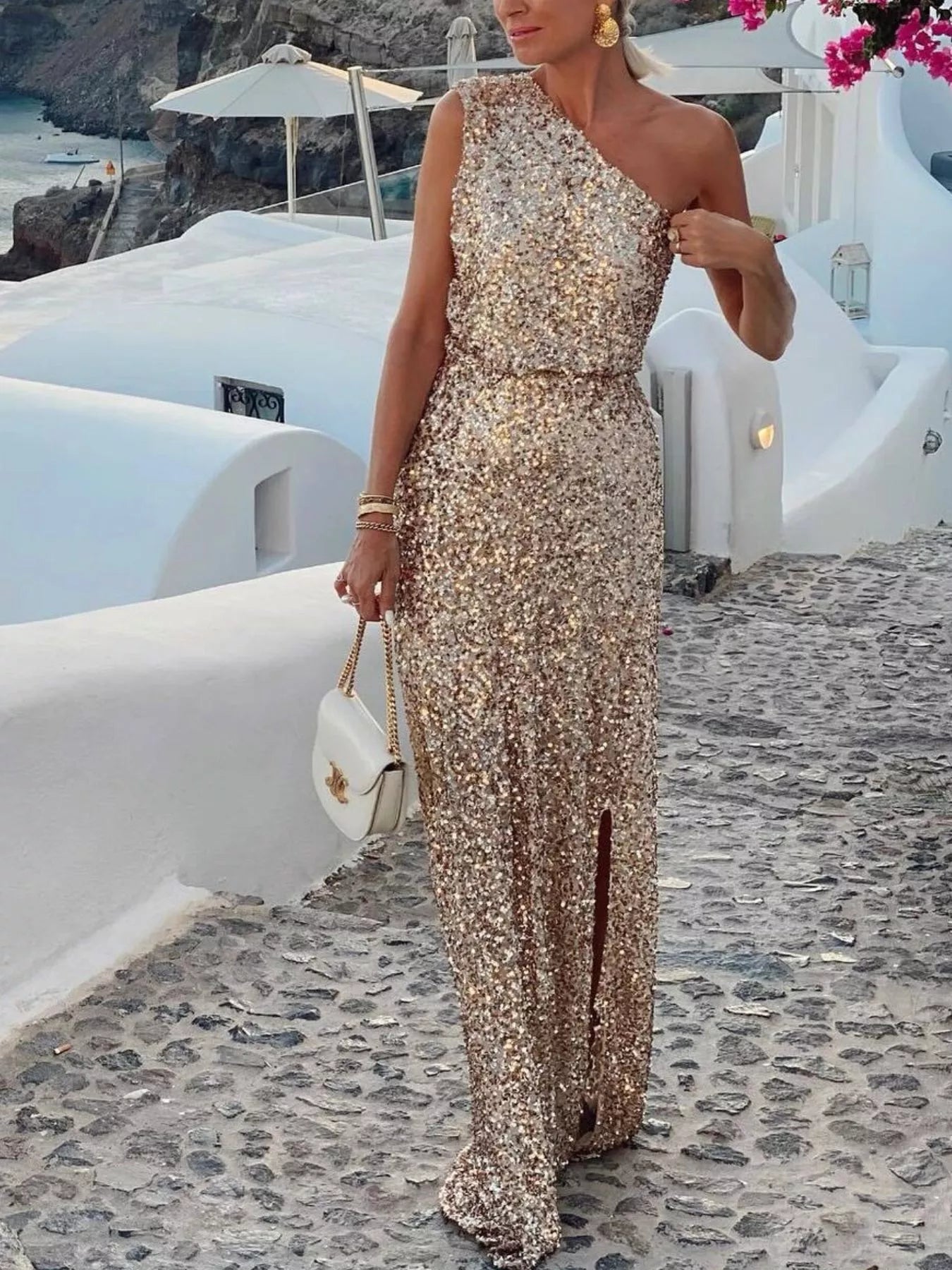 NEWYORKLISTED STYLE COLLECTION: Woman in a sparkly one-shoulder dress standing on a cobbled street with white buildings and blue sea in the background.