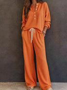 NEWYORKLISTED STYLE & COMFORT COLLECTION: A woman wearing a matching orange waffle-knit lounge set with a long-sleeve henley top and wide-leg pants, featuring a white drawstring, standing against a dark textured wall.
