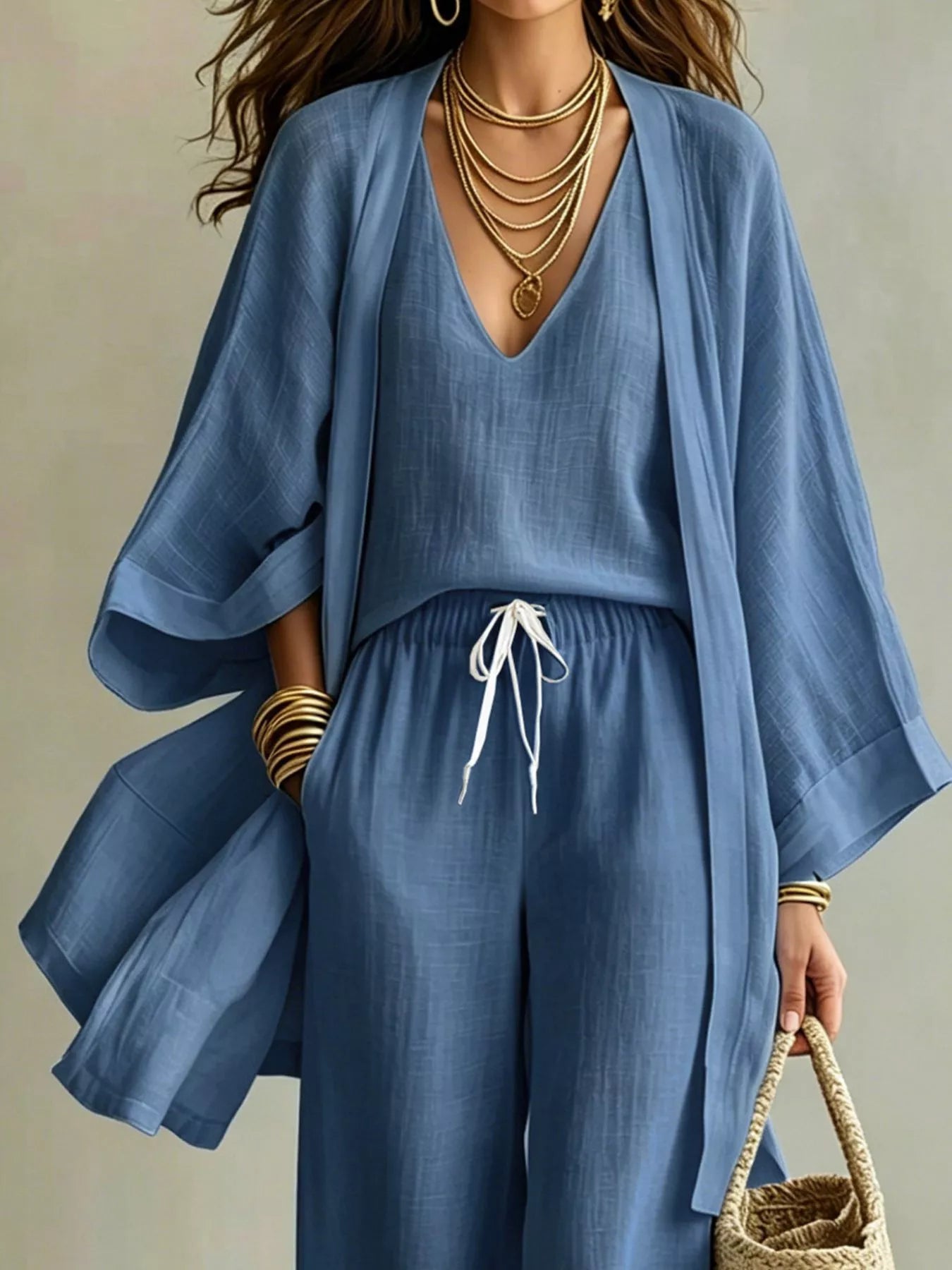 A woman wearing a matching blue NEWYORKLISTED STYLE COLLECTION: two-piece outfit with a loose long-sleeve top and wide-leg pants, styled with layered gold necklaces, bracelets, and a woven handbag against a neutral background.