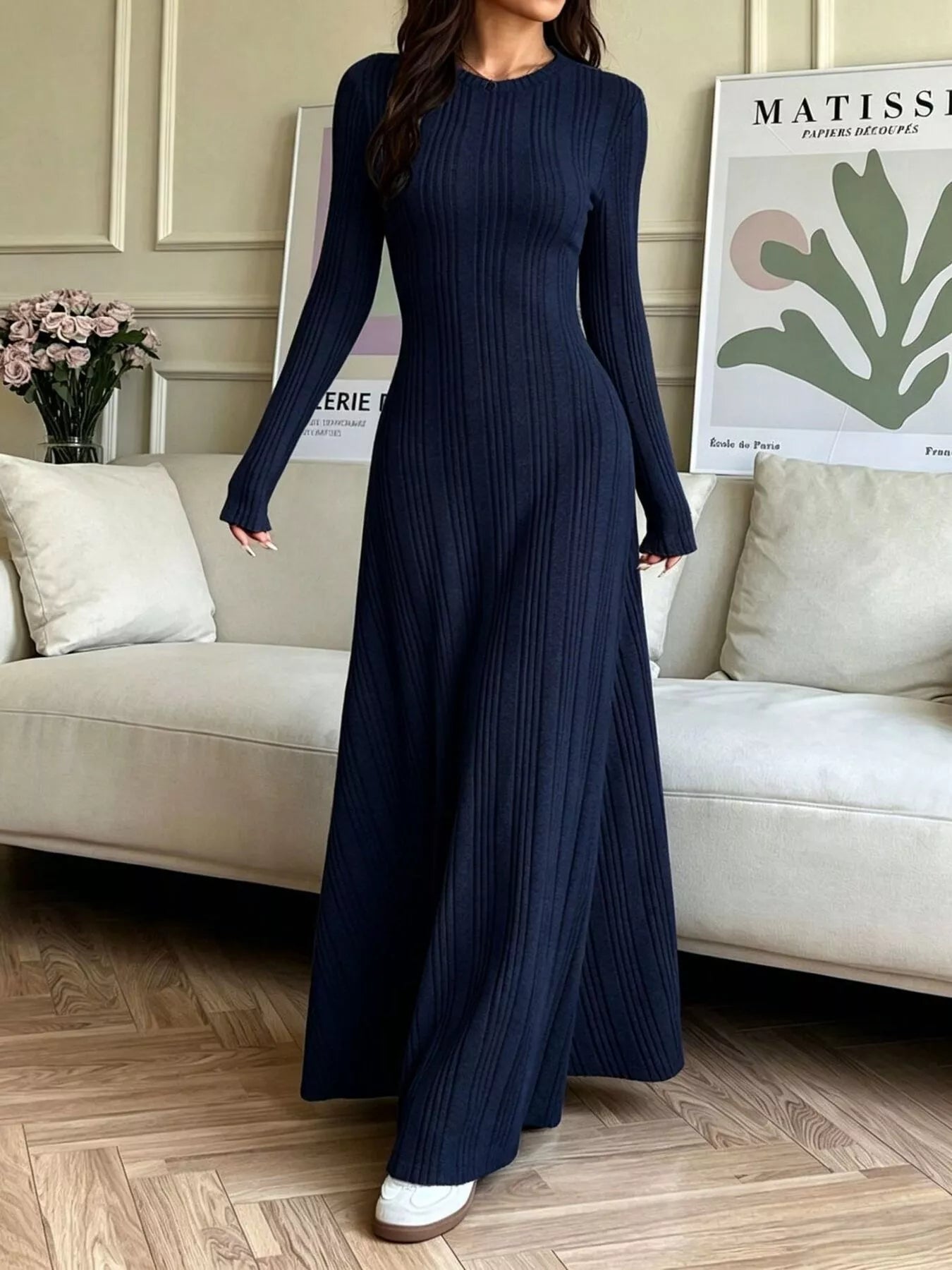 NEWYORKLISTED STYLE COLLECTION: A woman wearing a navy blue long-sleeve maxi dress with a vertical pleated texture, standing in a living room setting with a beige sofa and wooden floor.