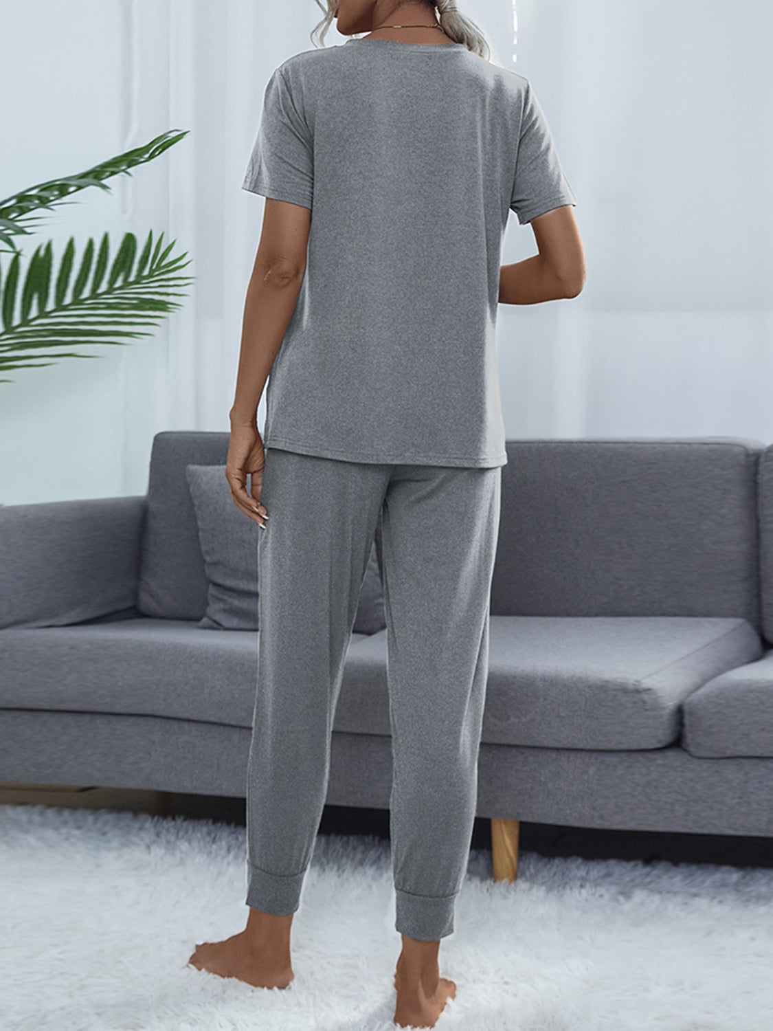 Person wearing gray pajamas standing in a living room with a gray sofa and white curtains.
