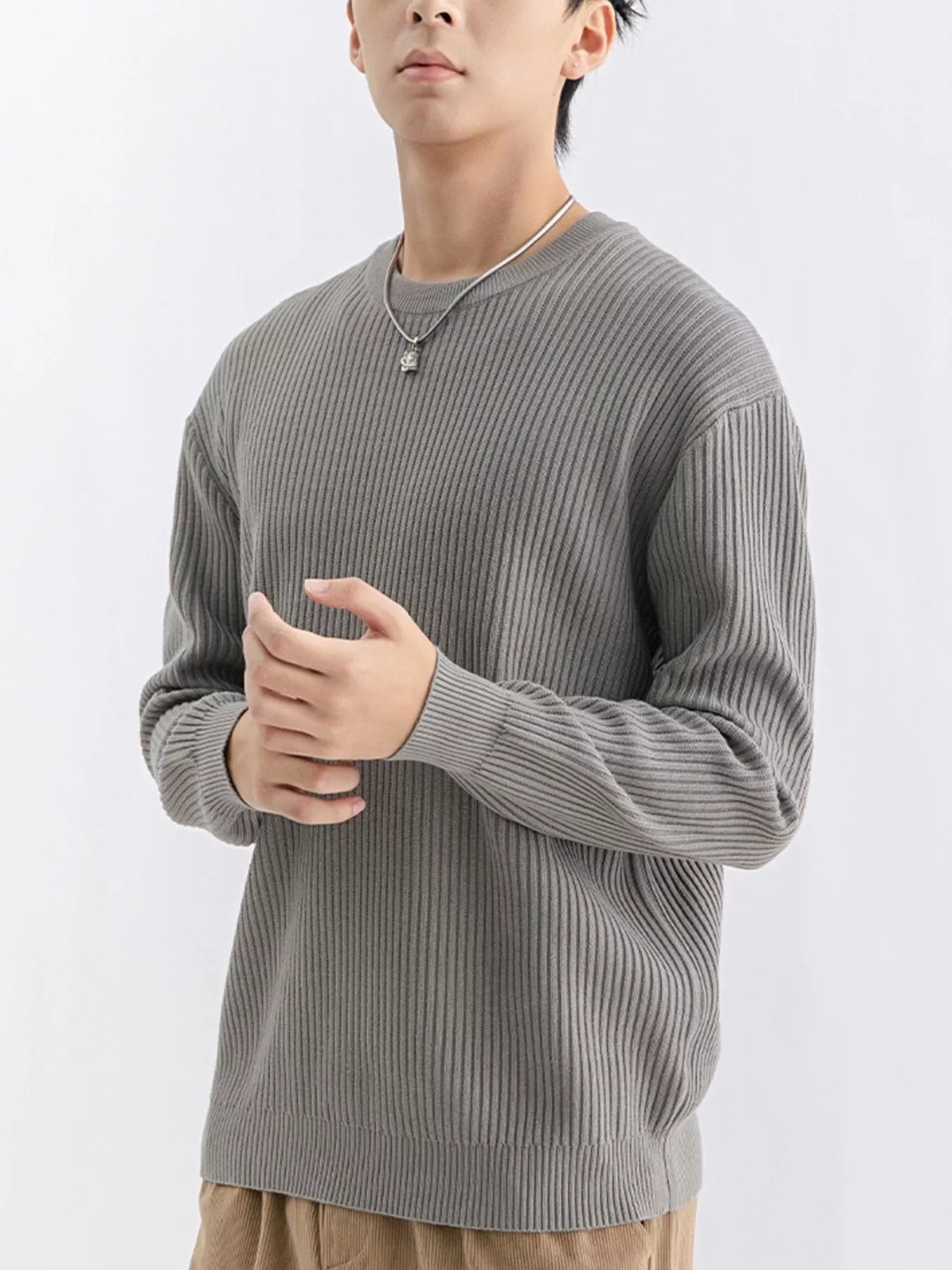 NEWYORKLISTED ATLAS MEN'S COLLECTION: A man wearing a ribbed gray long-sleeve crewneck sweater, paired with tan pants and a silver necklace, photographed against a clean white studio background.
