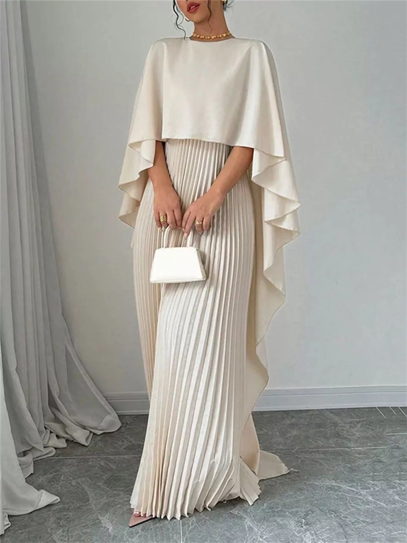 NEWYORKLISTED STYLE COLLECTION: Woman wearing a beige cape over a pleated dress holding a white clutch against a plain background