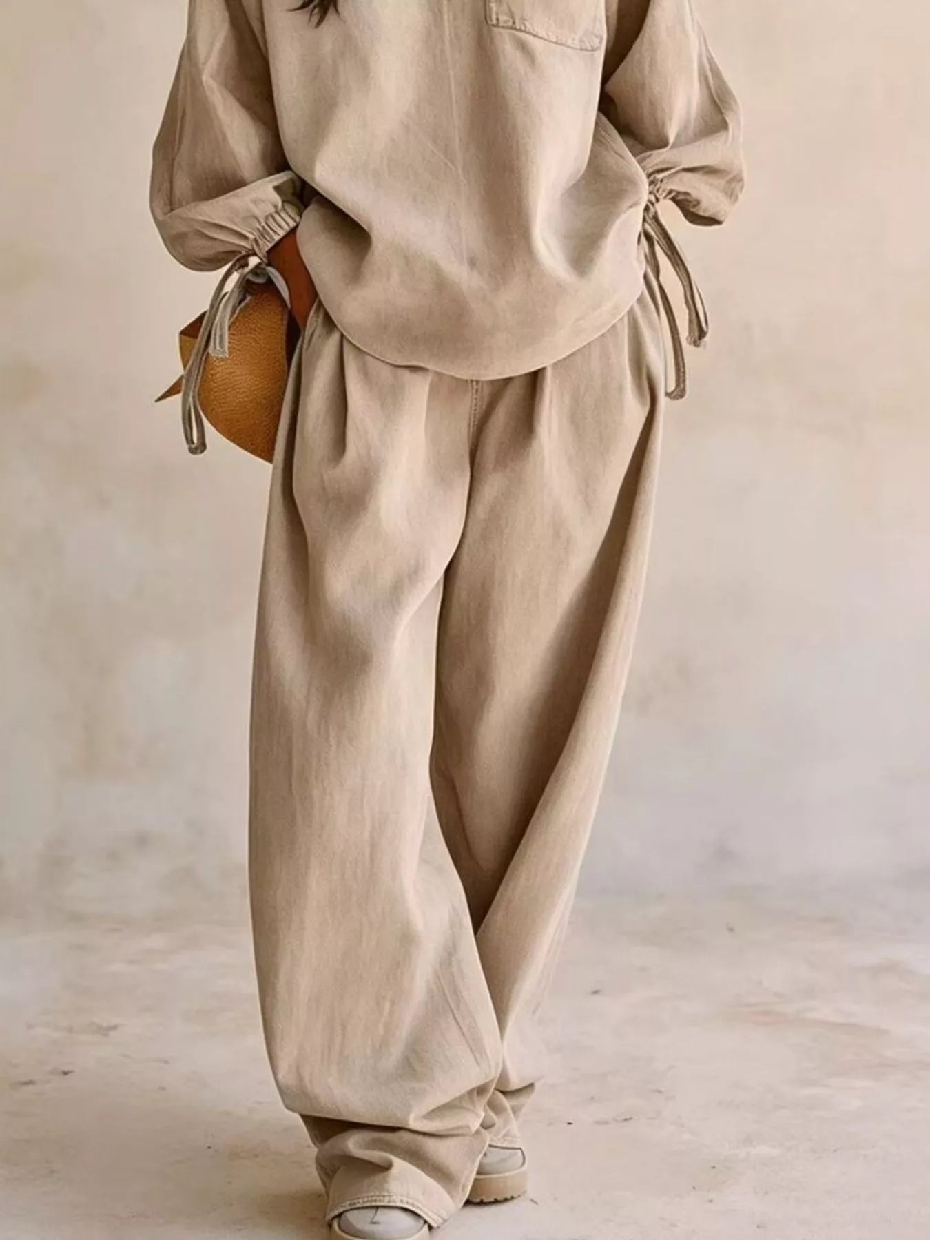 NEWYORKLISTED STYLE COLLECTION: A person wearing a matching beige linen-like two-piece outfit with a loose long-sleeve top and wide-leg pants, styled with a small round tan bag and white sneakers against a soft neutral wall.