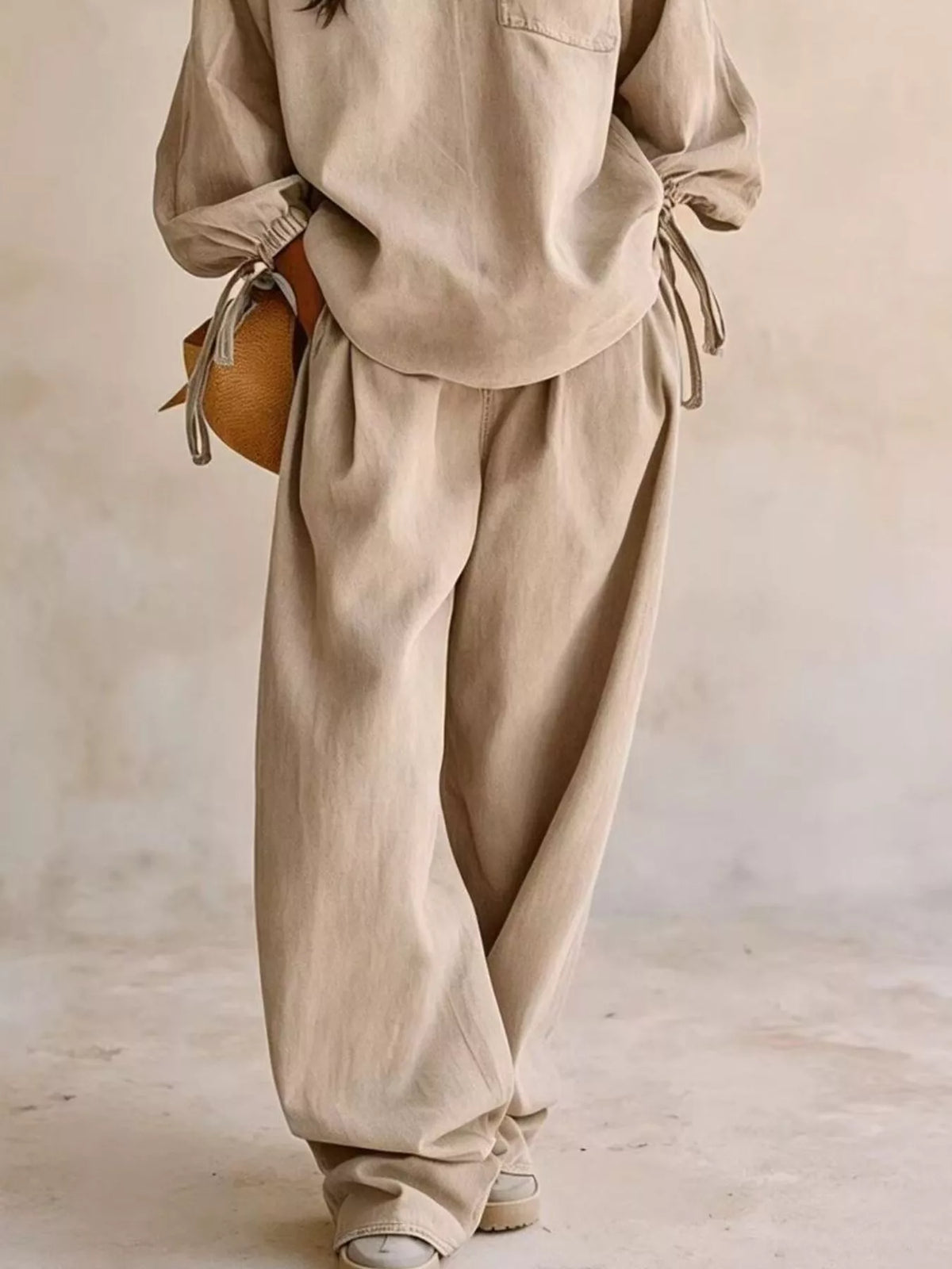 NEWYORKLISTED STYLE COLLECTION: A person wearing a matching beige linen-like two-piece outfit with a loose long-sleeve top and wide-leg pants, styled with a small round tan bag and white sneakers against a soft neutral wall.