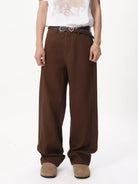 NEWYORKLISTED ATLAS MEN'S COLLECTION: A person wearing loose brown wide-leg pants with a dark belt and a small red heart-shaped buckle detail, paired with a white graphic T-shirt and tan shoes, photographed against a plain white studio background.