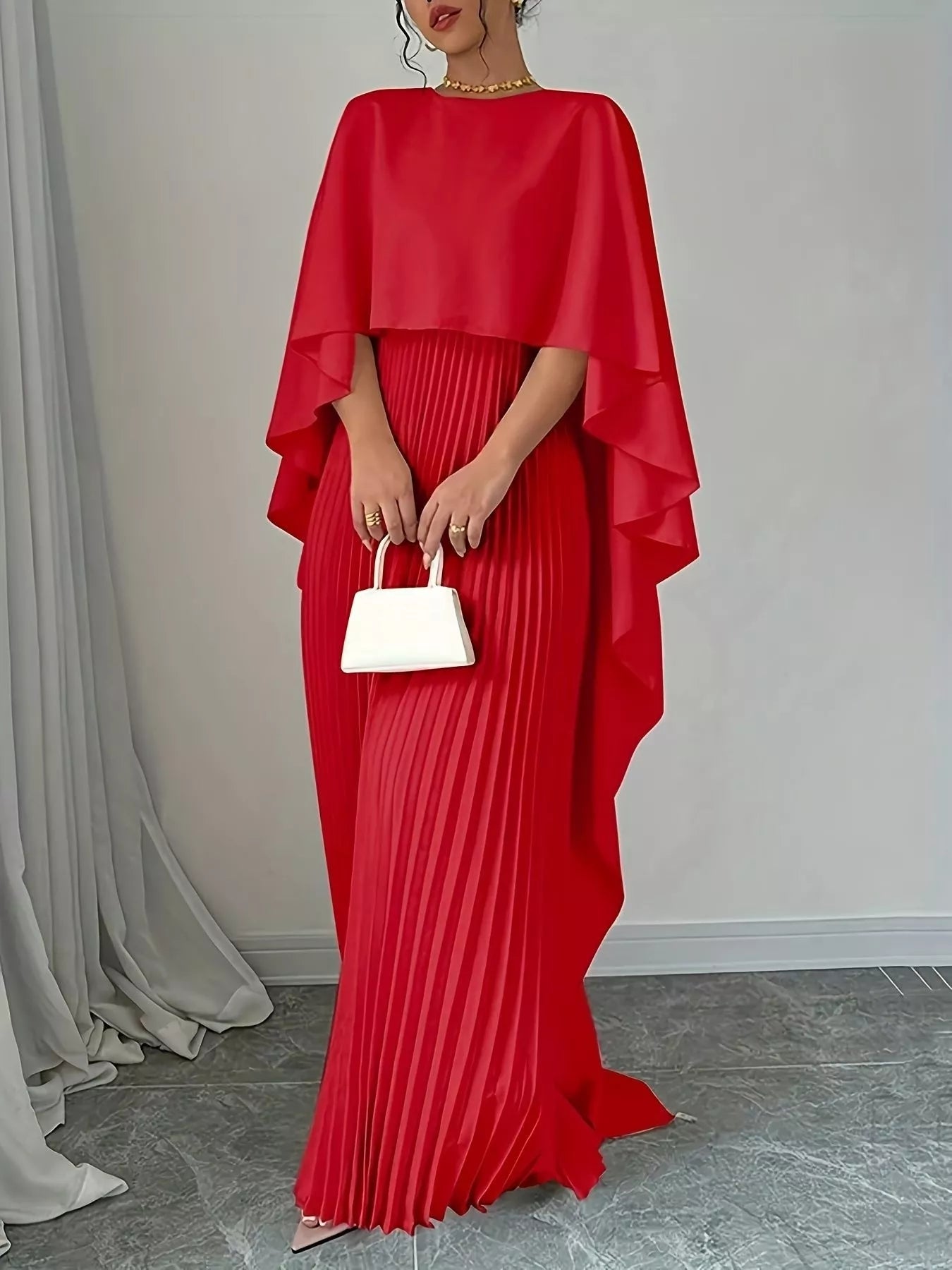 NEWYORKLISTED STYLE COLLECTION: Red pleated dress with cape held by a person against a neutral background
