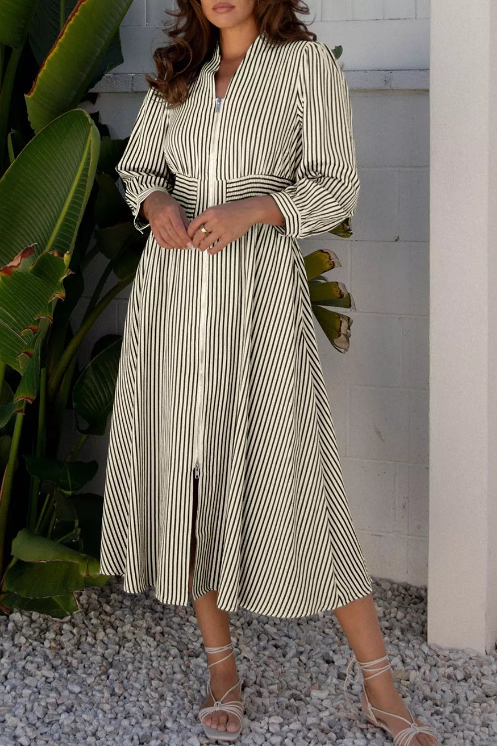 NEWYORKLISTED STYLE COLLECTION: Woman wearing a striped dress standing outdoors with plants and a white wall in the background