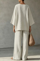 NEWYORKLISTED STYLE COLLECTION: A woman shown from the back wearing a loose off-white linen two-piece set with a wide-sleeve top and wide-leg trousers, holding a woven straw bag against a textured gray wall.