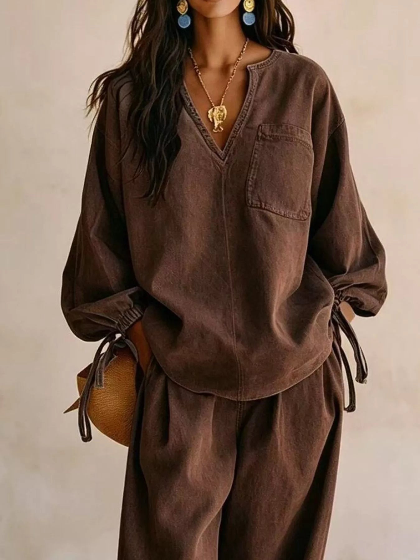 NEWYORKLISTED STYLE COLLECTION: A woman wearing a loose brown long-sleeve top and matching wide-leg pants, styled with a gold necklace, blue earrings, and a brown woven bag against a neutral beige background.