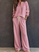 NEWYORKLISTED STYLE & COMFORT COLLECTION: A woman wearing a matching dusty pink lounge set consisting of a long-sleeve button-front top and wide-leg pants with a cream drawstring, standing against a dark textured wall.