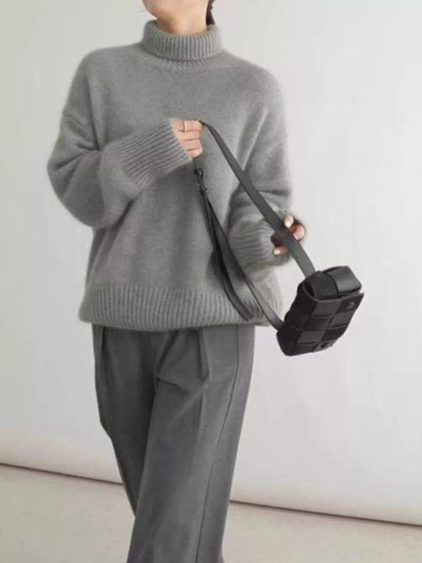 Person wearing a gray sweater and pants holding a black bag against a white background