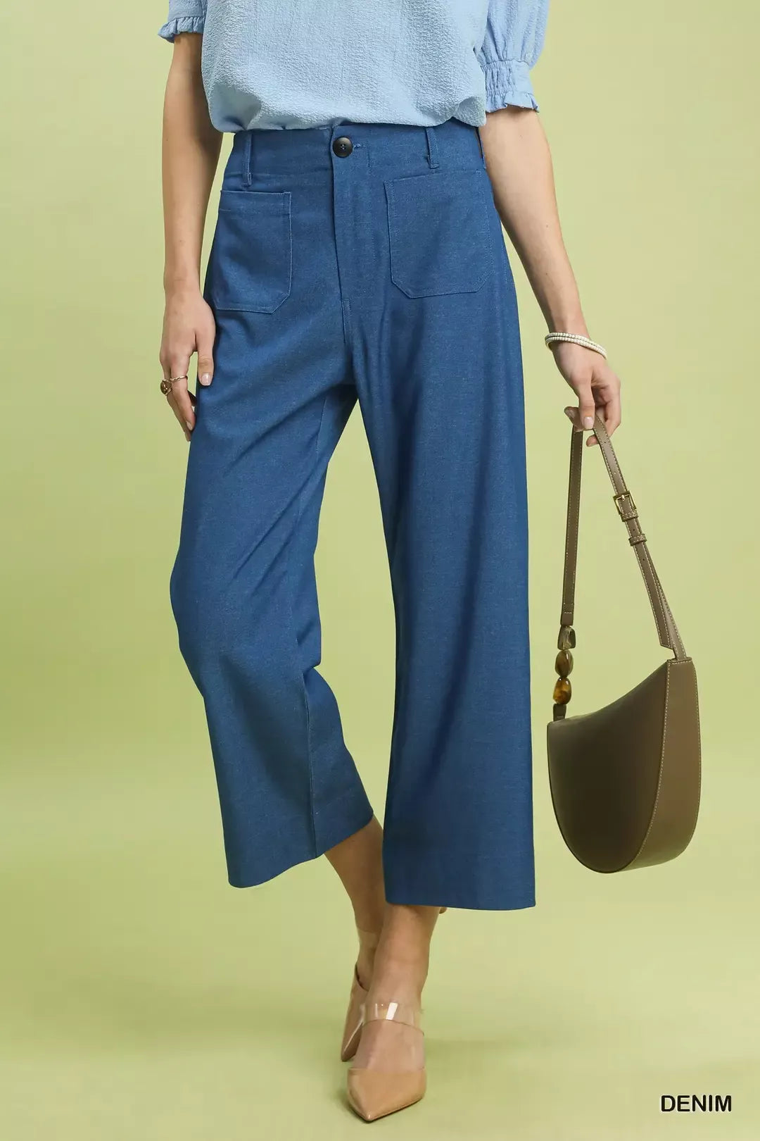 NEWYORKLISTED Style Collection: Person wearing blue pants and a light blue shirt, holding a brown handbag on a green background.