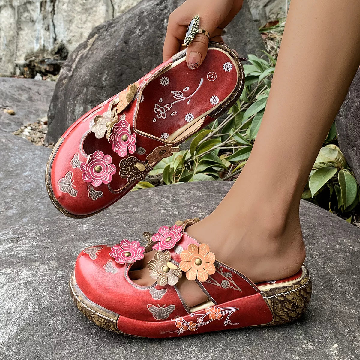 NEWYORKLISTED SHOE COLLECTION: Red floral sandals being worn on a stone surface with greenery in the background