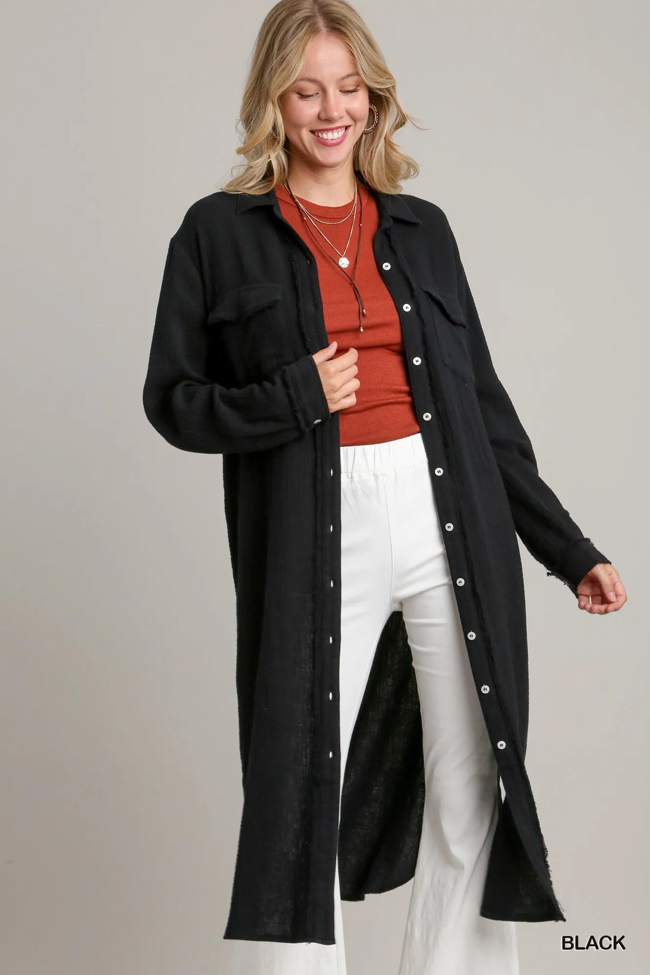 NEWYORKLISTED STYLE COLLECTION: Woman wearing a black long coat over a red top and white pants on a gray background