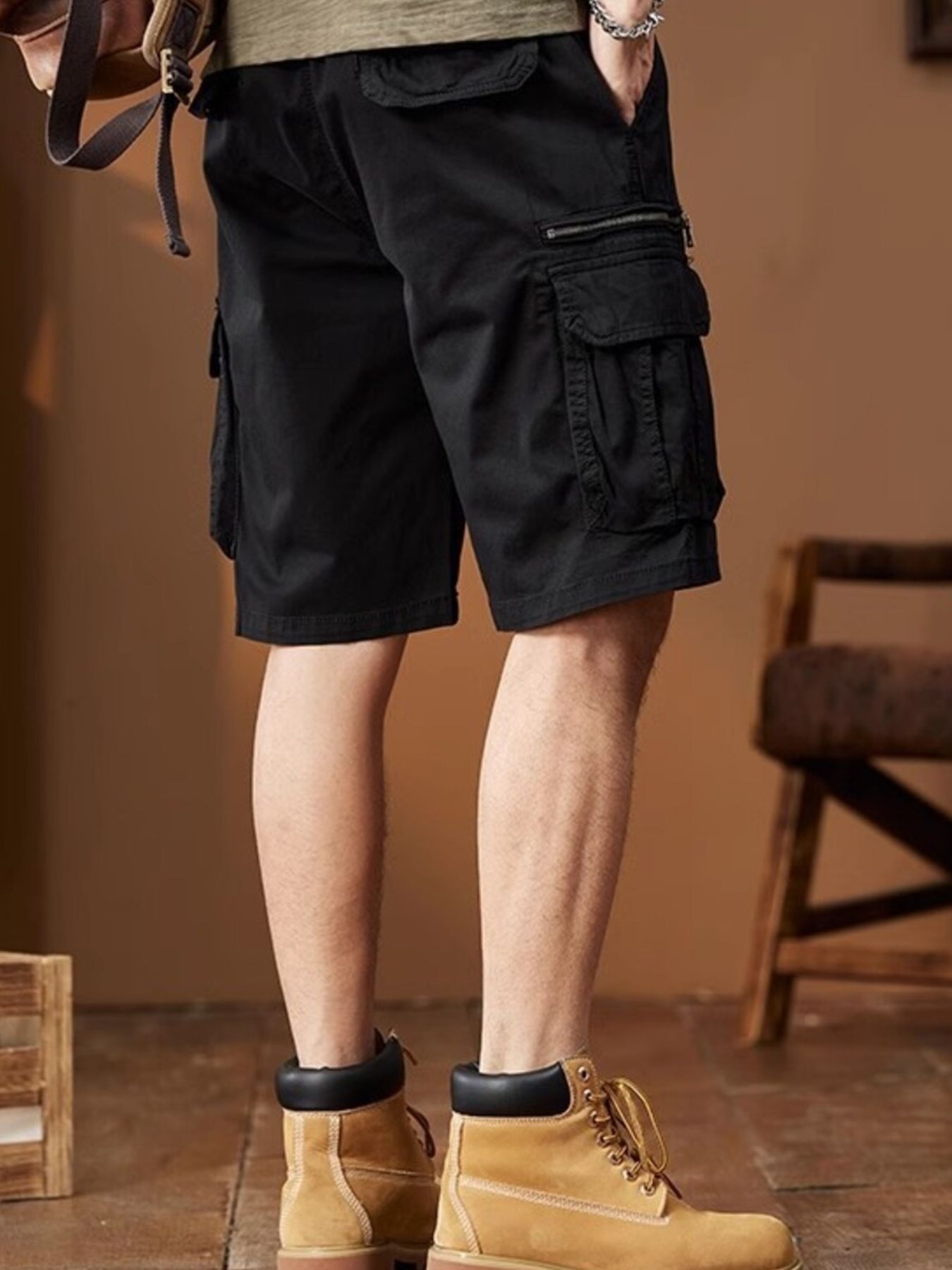 NEWYORKLISTED ATLAS MEN's COLLECTION: Person wearing black cargo shorts and tan boots in a room with a wooden floor and chair.
