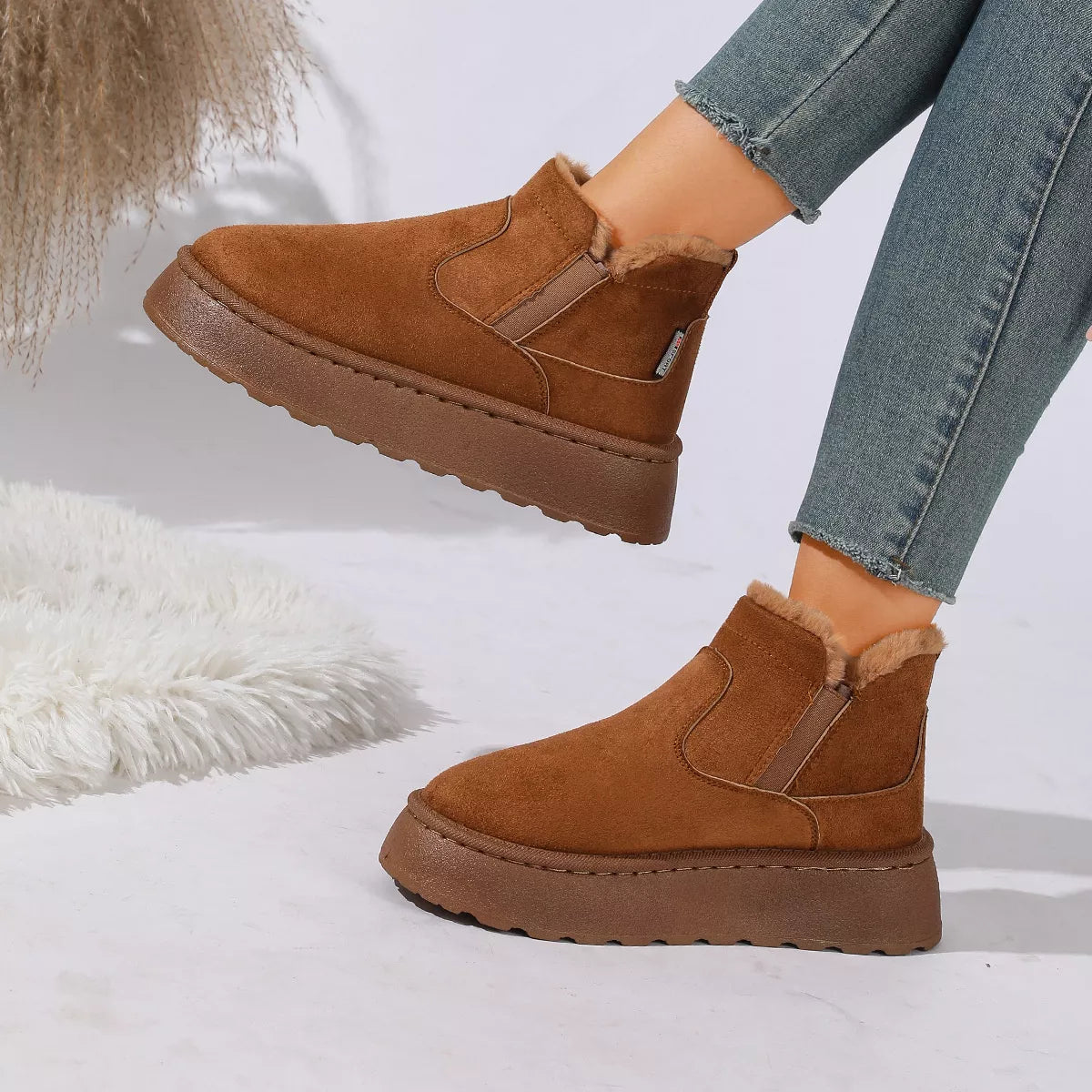NEWYORKLISTED SHOE COLLECTION: A pair of women's brown suede ankle boots with a plush faux-fur lining and chunky platform soles, shown on a model wearing blue jeans against a white studio background with decorative faux fur.