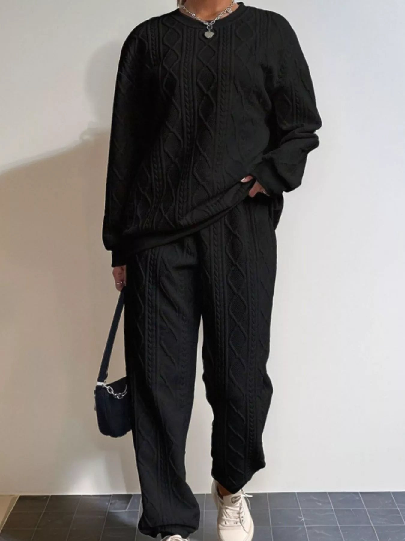 NEWYORKLISTED COMFORT COLLECTION: Person wearing a black cable knit sweater and pants with a white wall background